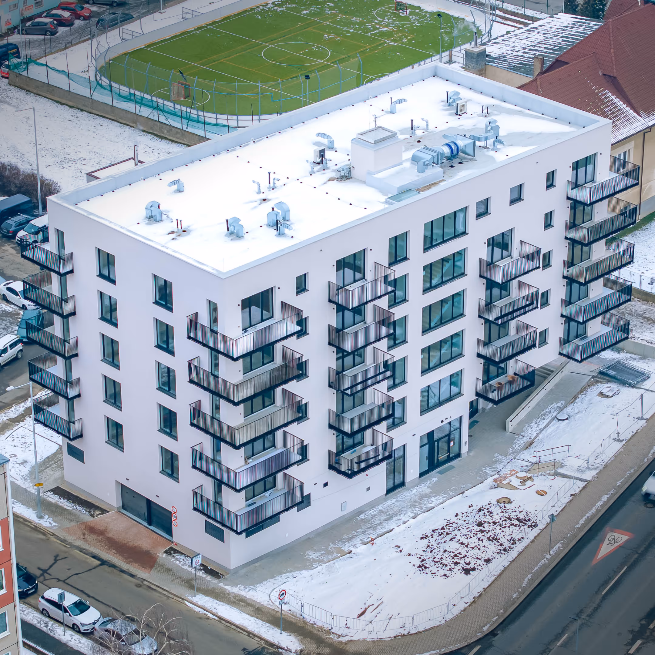 Nový bytový dům se sněhem na střeše a kolem něj, s balkóny a za ním hřiště s fotbalovým polem.
