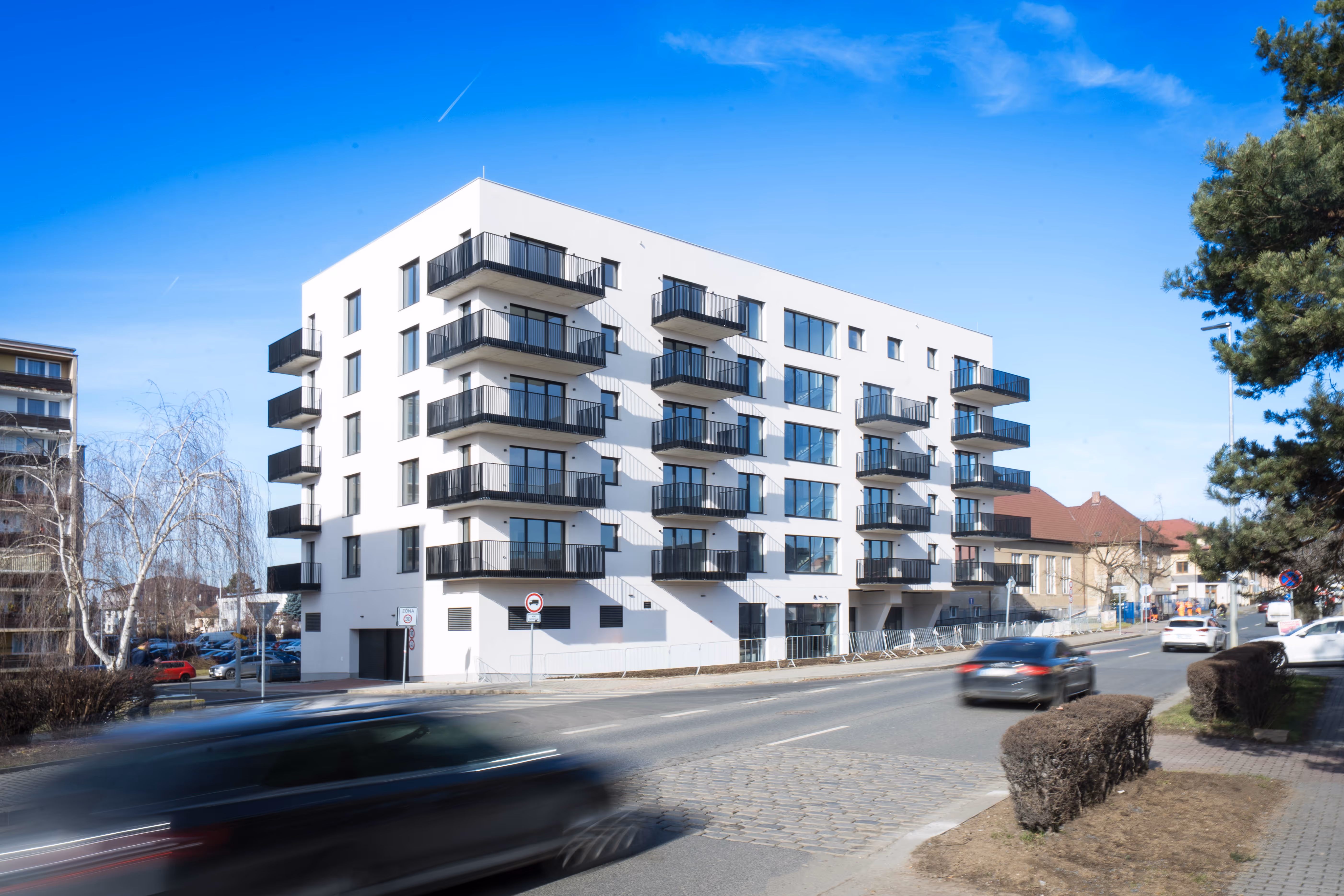 Moderní bílá bytová budova s balkóny v zimním městském prostředí s okolními paneláky a sněhem na zemi.