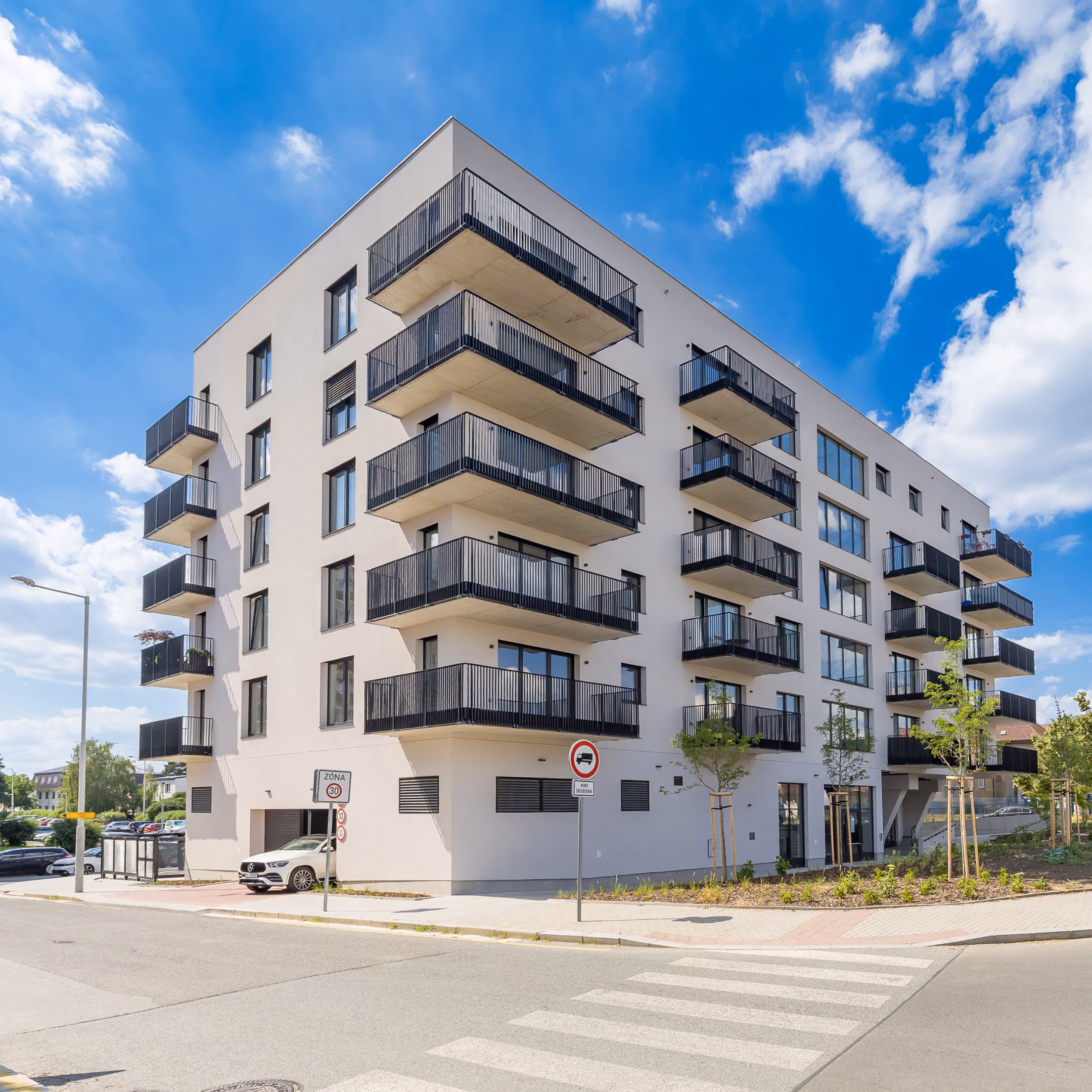 Moderní pětipodlažní bytový dům s balkony a přechodem pro chodce v popředí.