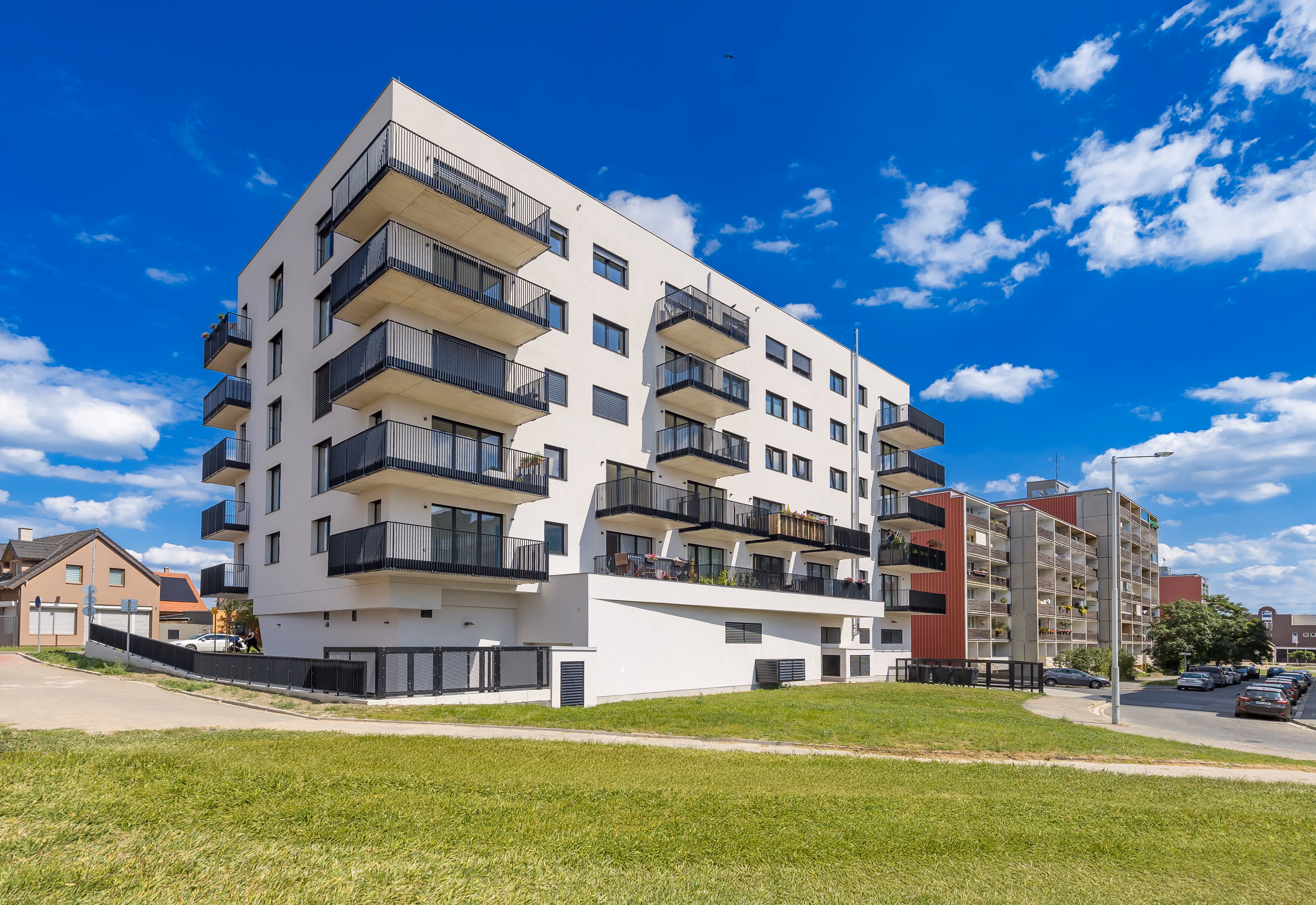Moderní bytový dům s balkony a zeleným prostorem na slunečný den s modrou oblohou.