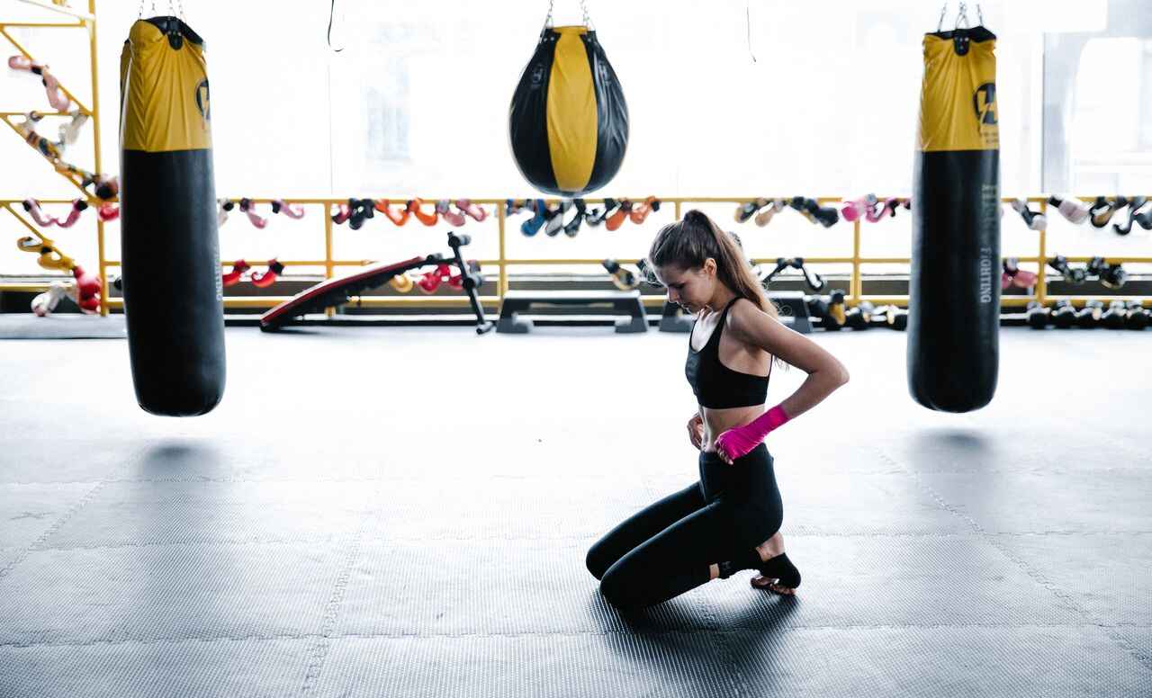 Woman in fitness attire kneeling in a gym beside punching bags, representing healthy lifestyle habits that support weight loss goals.