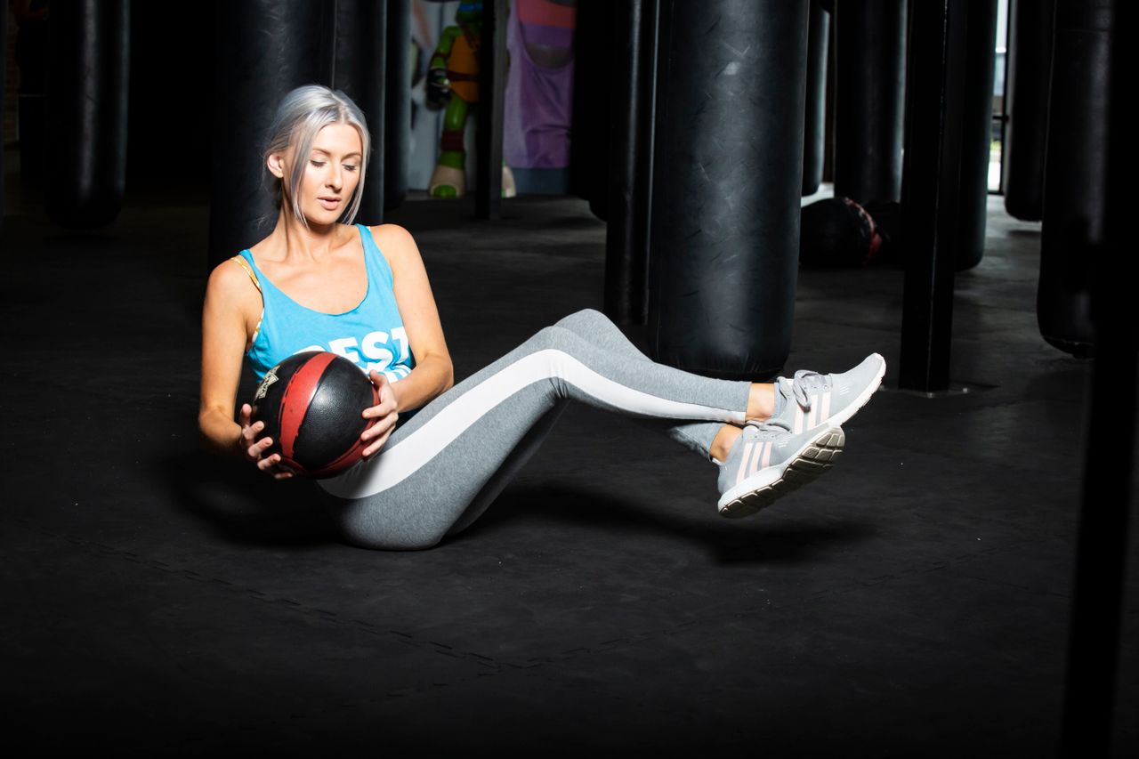 Woman performing a seated Russian twist with a medicine ball in a gym, demonstrating a core-strengthening exercise for balance, stability, and fitness.