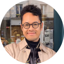 Man with glasses and short curly hair wearing a beige jacket and black turtleneck standing in front of a café window.