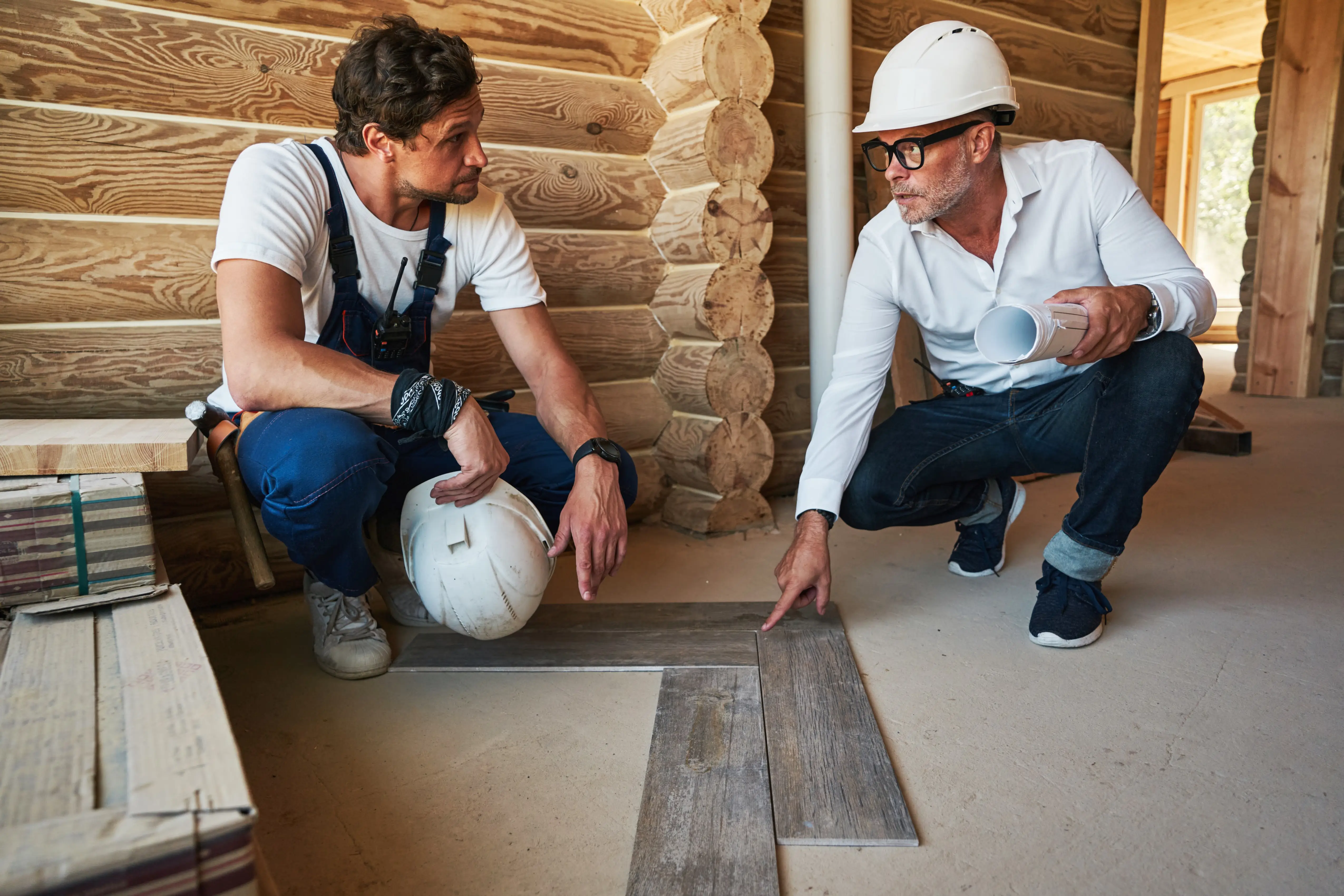 A contractor in overalls and a project manager holding blueprints crouch down together inside a log-frame home under construction, examining a wood-look plank flooring sample laid on a concrete subfloor
