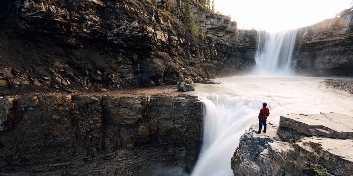 Person in a red plaid shirt stands on a rocky ledge by twin waterfalls in Clearwater Country.