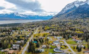 Aerial view of a small mountain town with forested streets, a lake, and snow-capped peaks under cloudy skies.