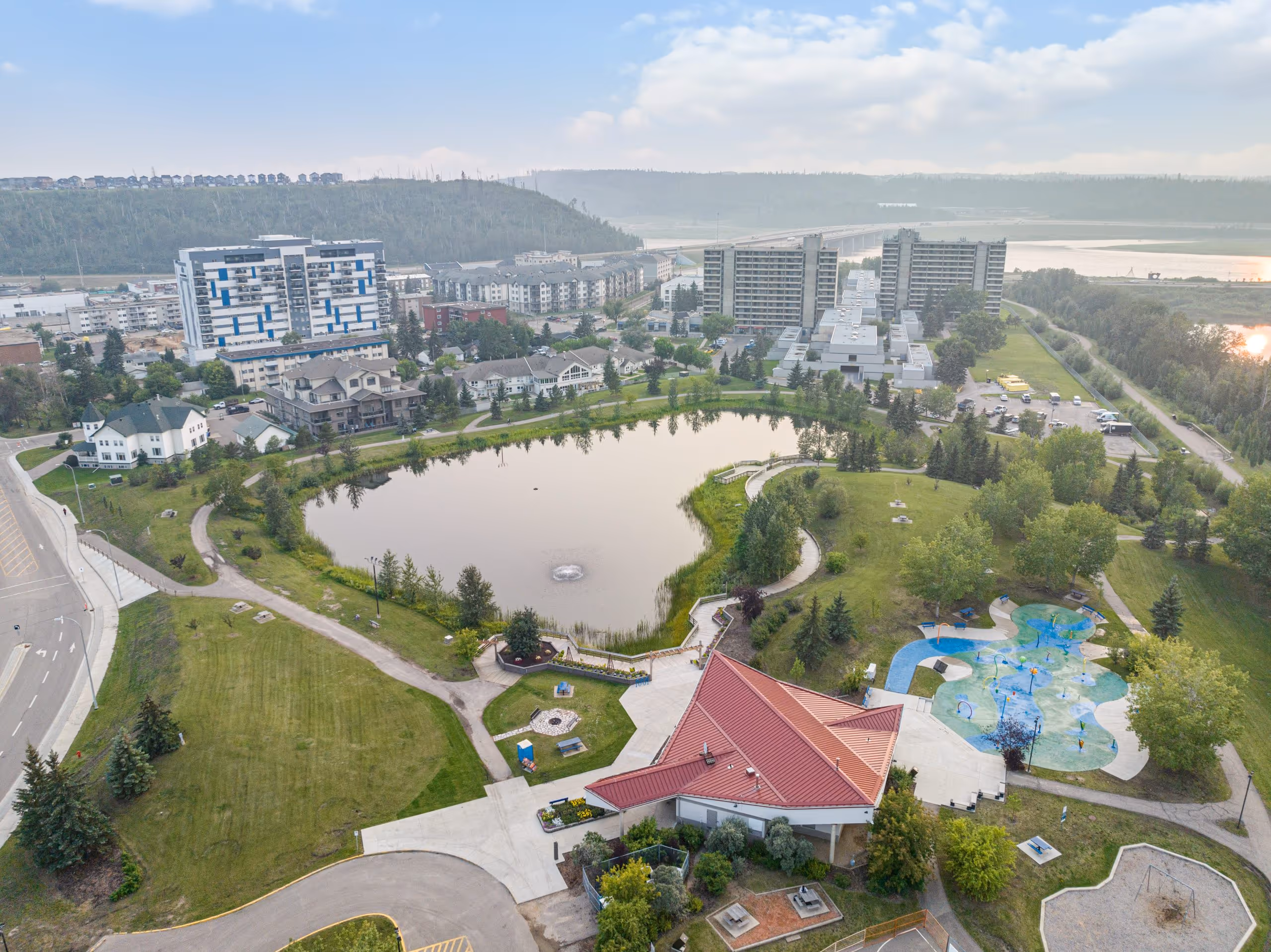 Aerial view of a park with pond and splash pad in Wood Buffalo, with apartments and a river in the background.