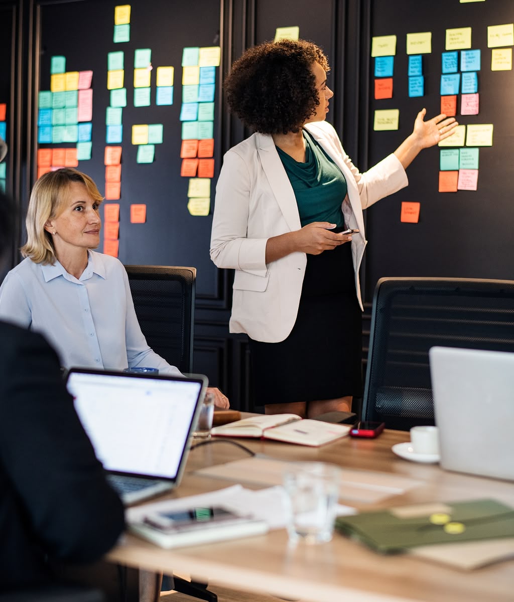 Business team in a meeting, woman presenting strategy on a wall covered with colorful sticky notes, colleagues listening and taking notes.