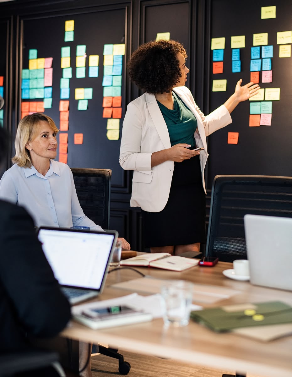 Business team in a meeting, woman presenting strategy on a wall covered with colorful sticky notes, colleagues listening and taking notes.