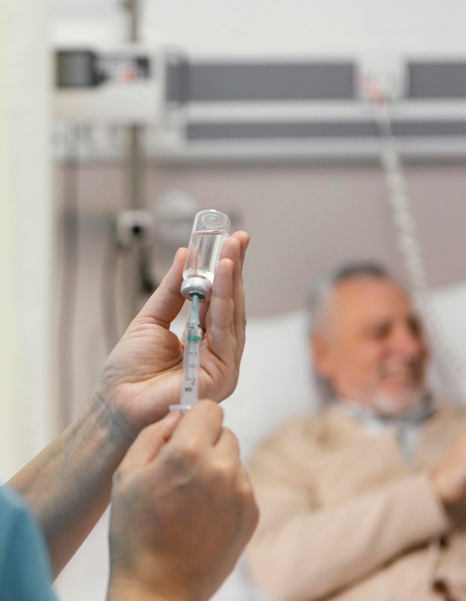Healthcare worker preparing a syringe for infusion therapy while a patient rests in a hospital bed in the background.