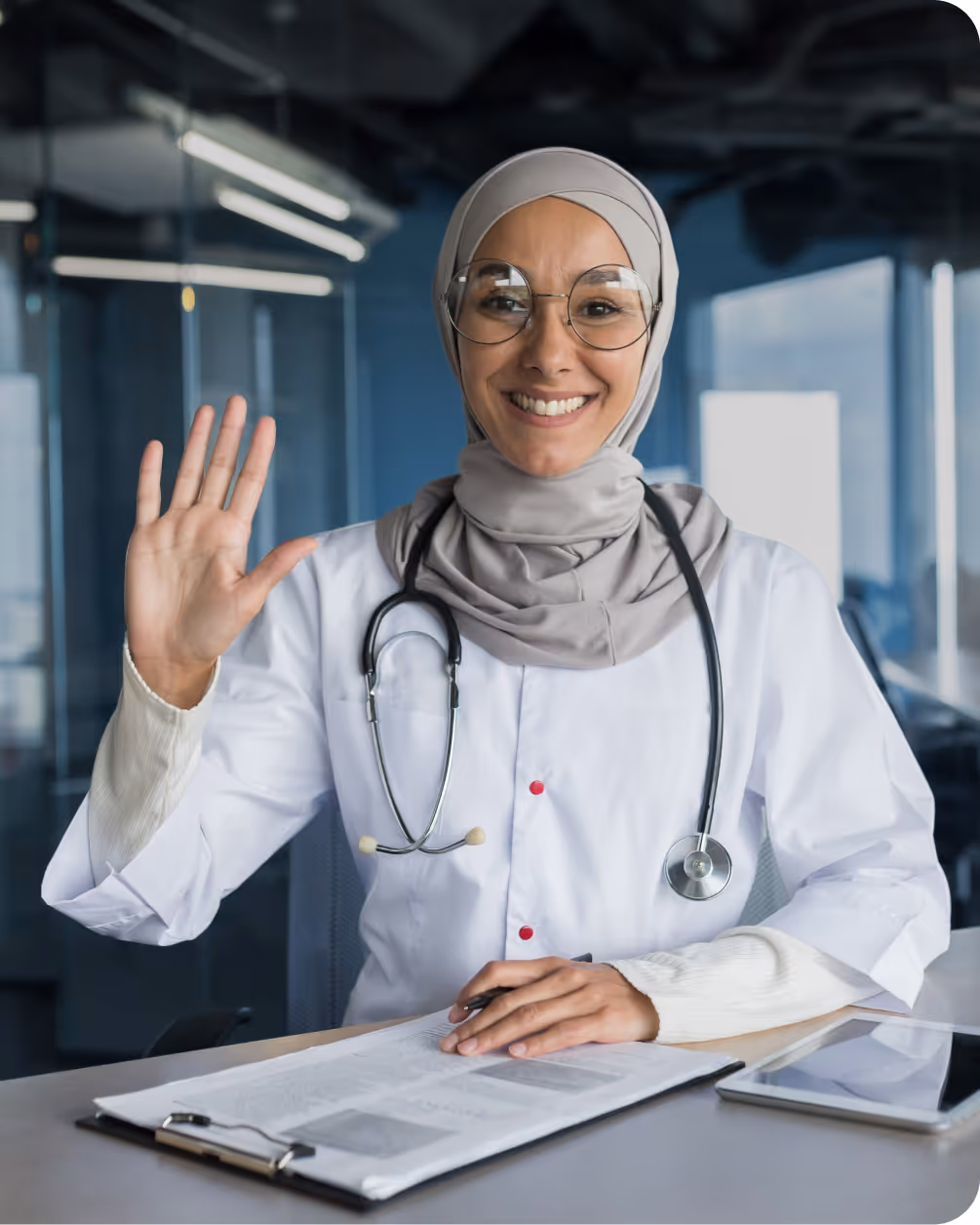 Smiling female doctor wearing a hijab and glasses, sitting at a desk with a clipboard and tablet, waving with her right hand.