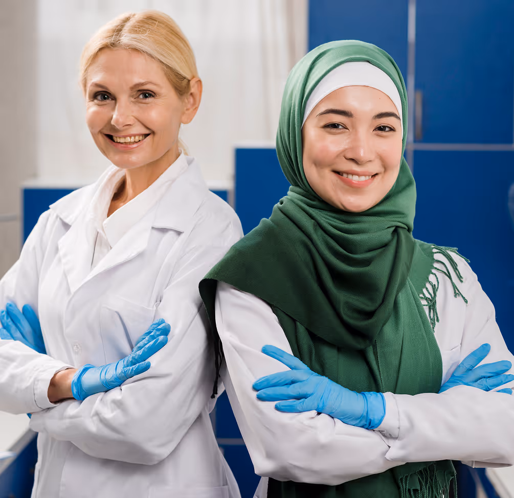 Two female scientists wearing lab coats and blue gloves, one with a green hijab, smiling confidently with arms crossed.