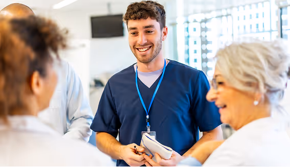 Smiling male healthcare professional in navy scrubs holding a notepad and talking to three colleagues in a bright medical setting.