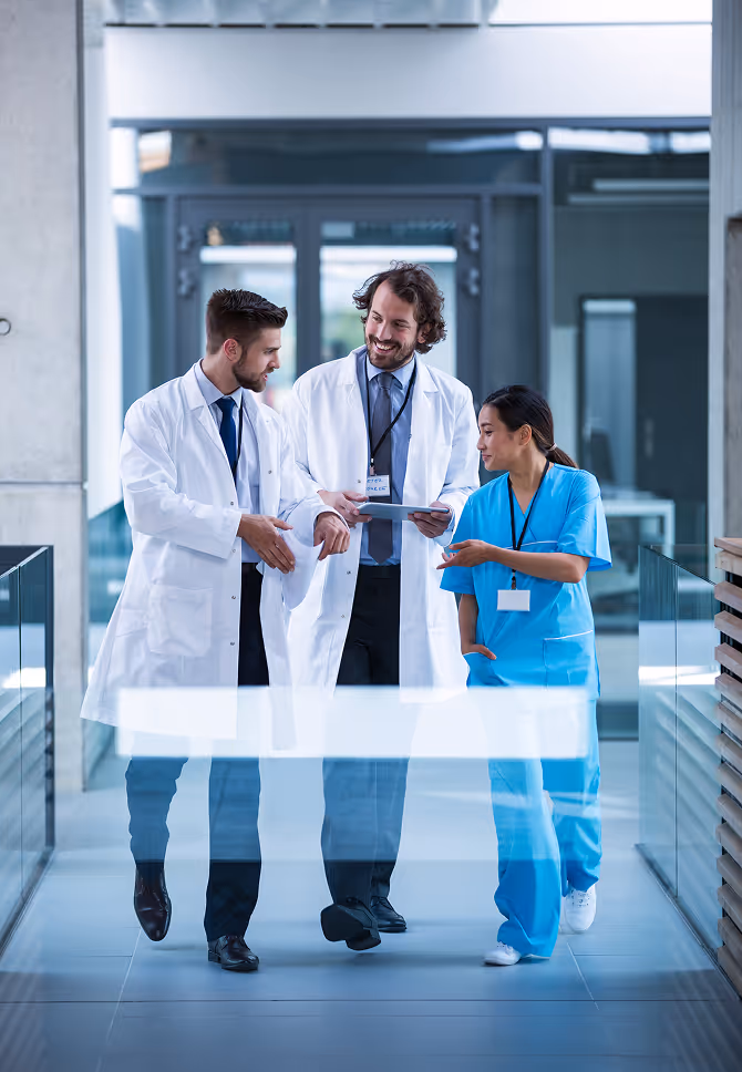 Two male doctors in white coats and a female nurse in blue scrubs walking and discussing in a hospital corridor.