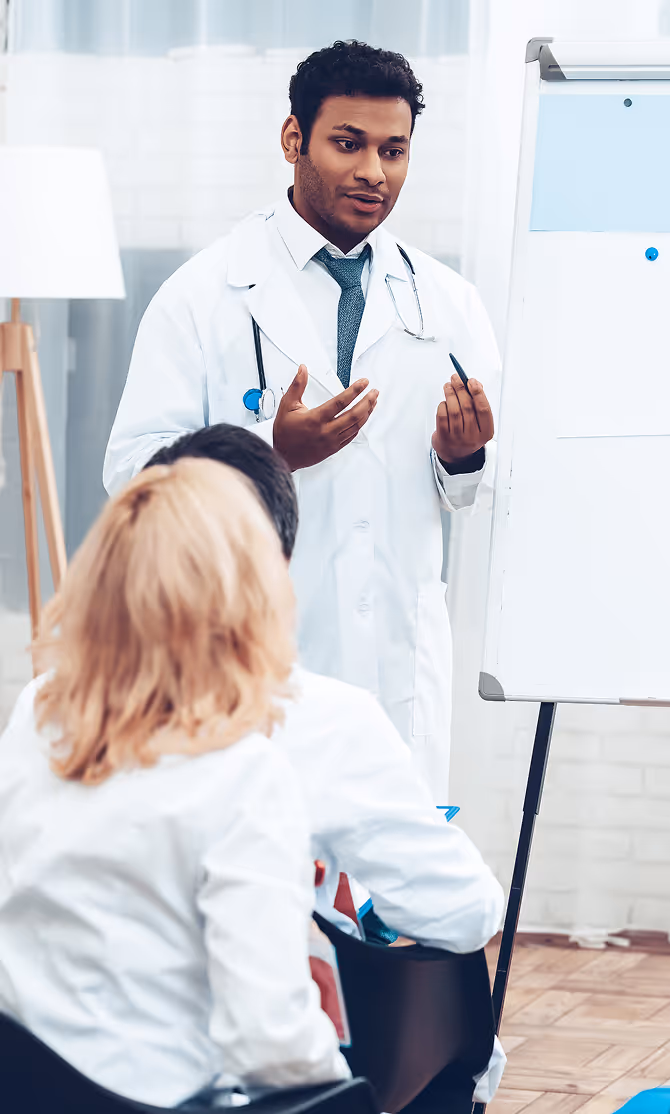 Male doctor in a white coat giving a presentation to seated colleagues in a bright room with a flip chart.