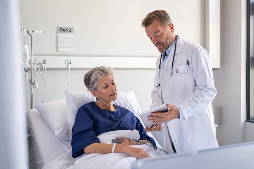 Doctor in white coat with stethoscope showing a tablet to an elderly female patient lying in a hospital bed labeled Bed 2.