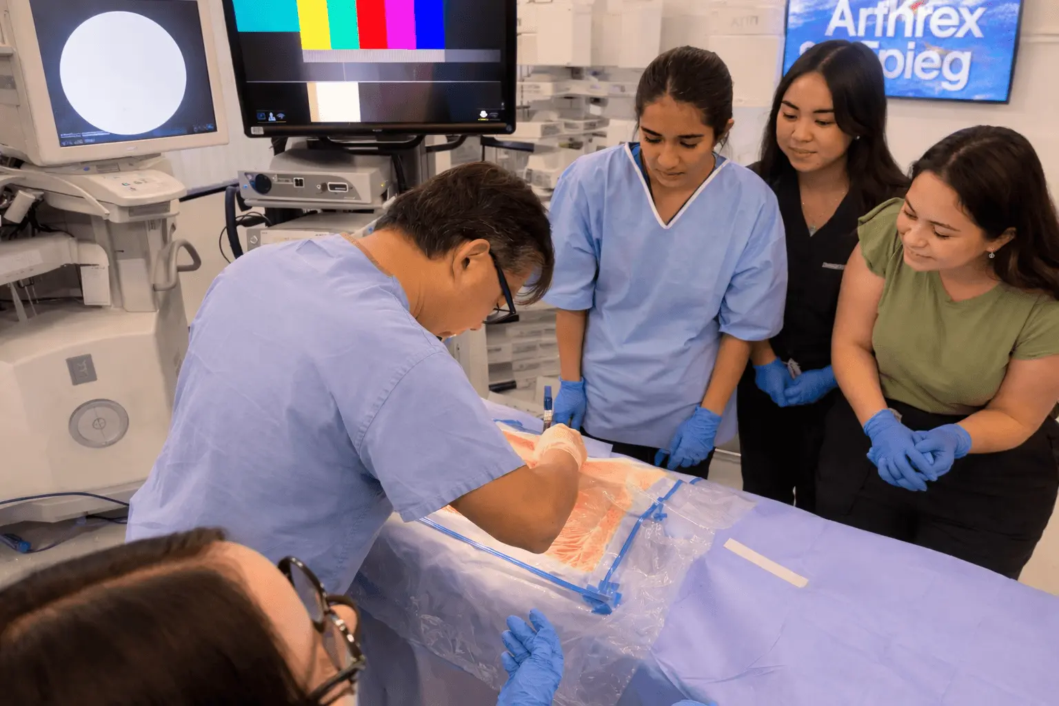Medical instructor demonstrates procedure on a model while three students watch closely wearing gloves.