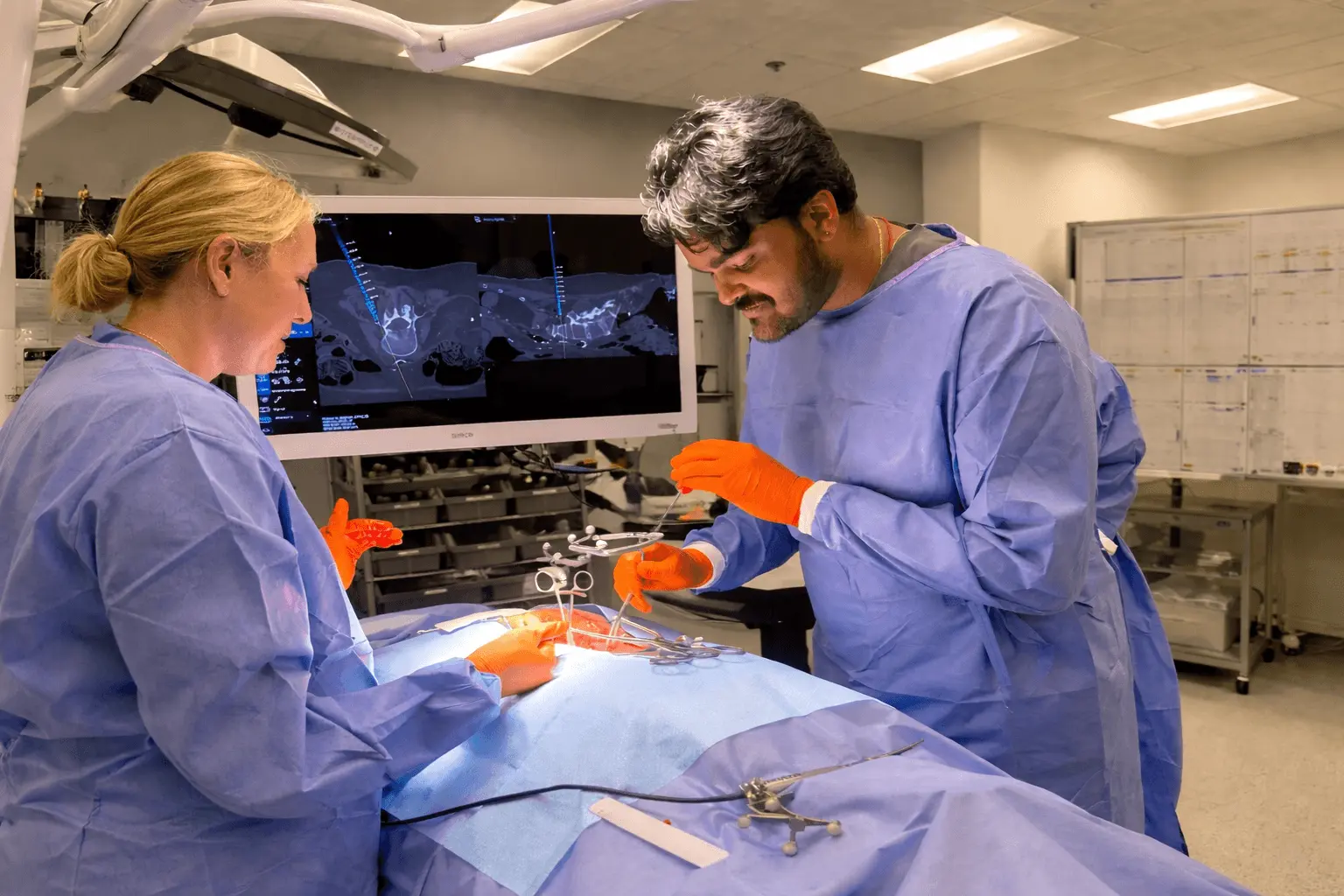 Two surgeons in blue gowns and orange gloves performing surgery with a medical scan on a screen behind them.