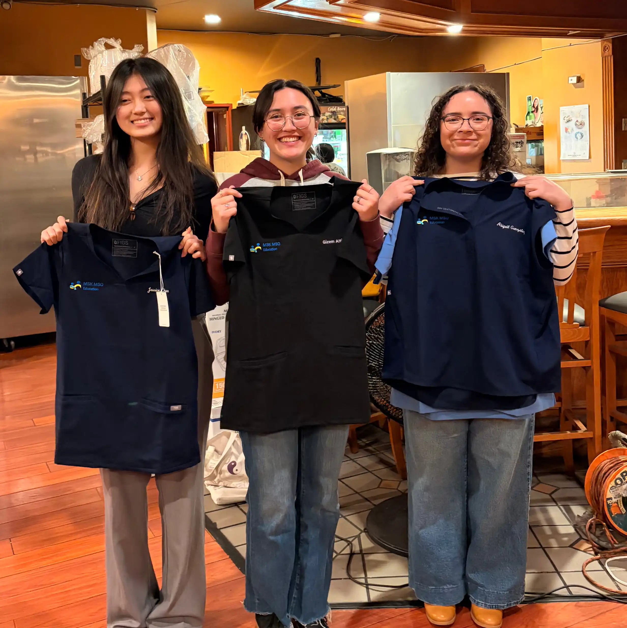 Three women standing indoors holding navy and black medical scrub tops with embroidered names.