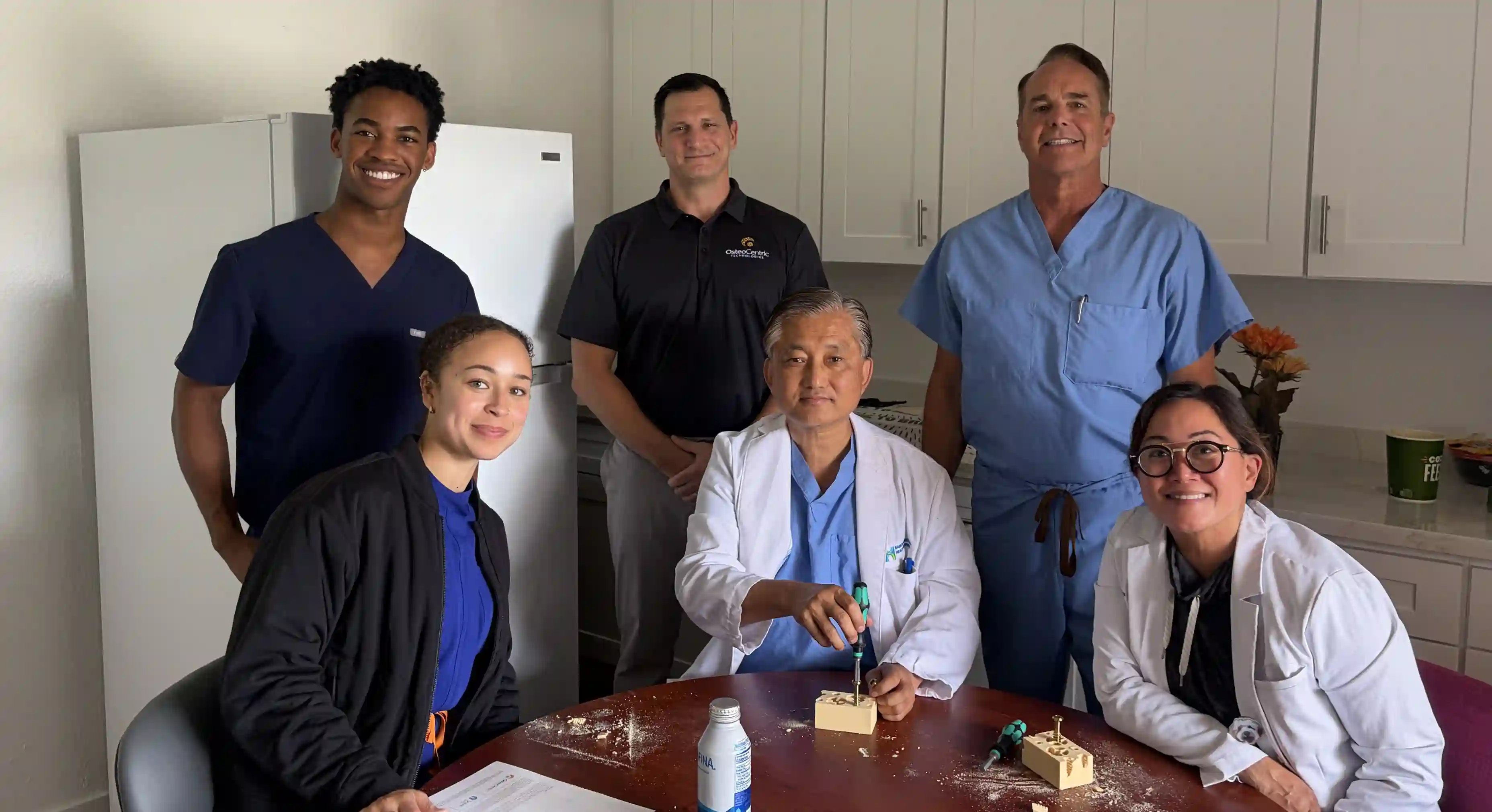 Five medical professionals and Dr. Yoo smiling, two seated at a table with bone models and screwdrivers in a clinical setting.