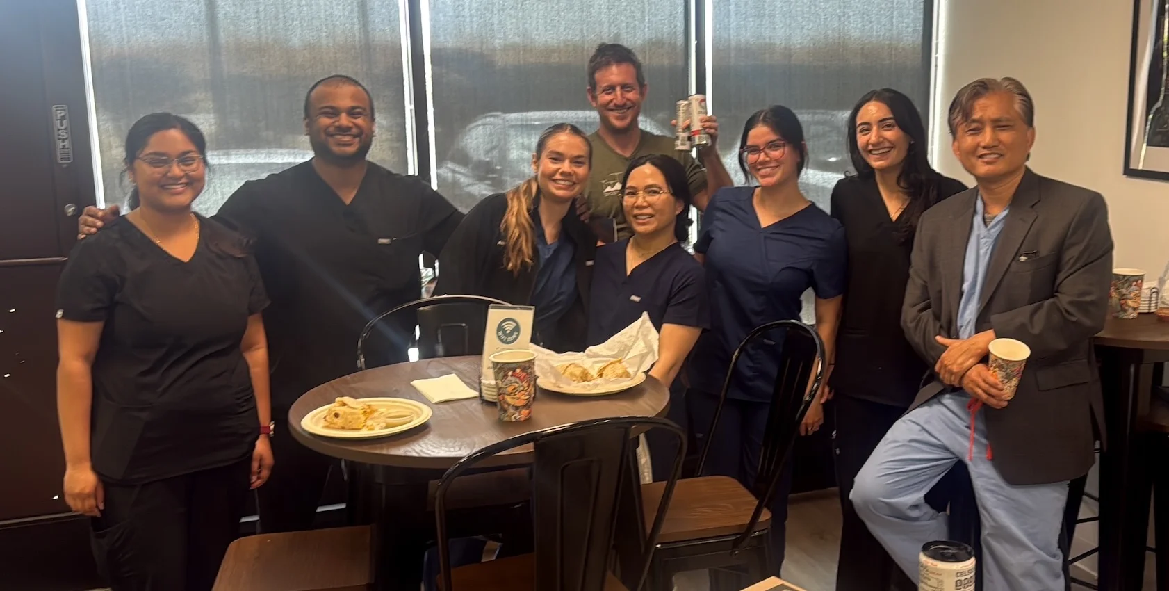 Group of seven smiling students and Dr Yoo in scrubs and casual clothes standing around a table with food and drinks.