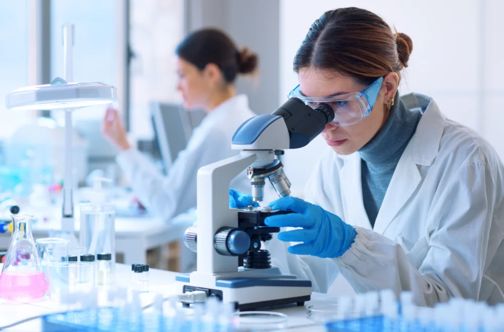 Female scientist wearing gloves and goggles looking through a microscope in a laboratory.