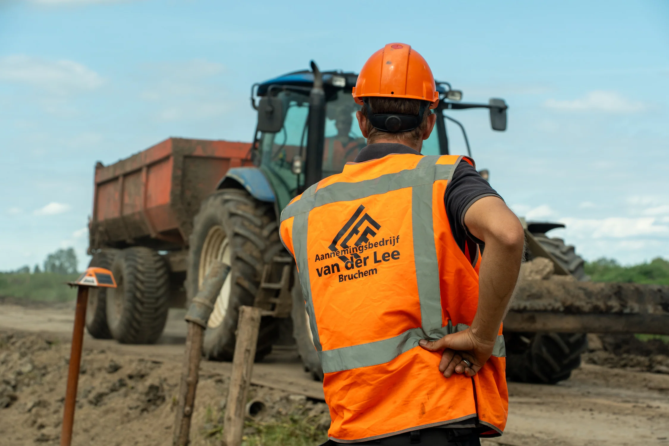 Een medewerker van Van der Lee Bruchem kijkt toe terwijl een tractor met aanhanger voorbijrijdt op het werkterrein tijdens dijkverzwaringswerkzaamheden.