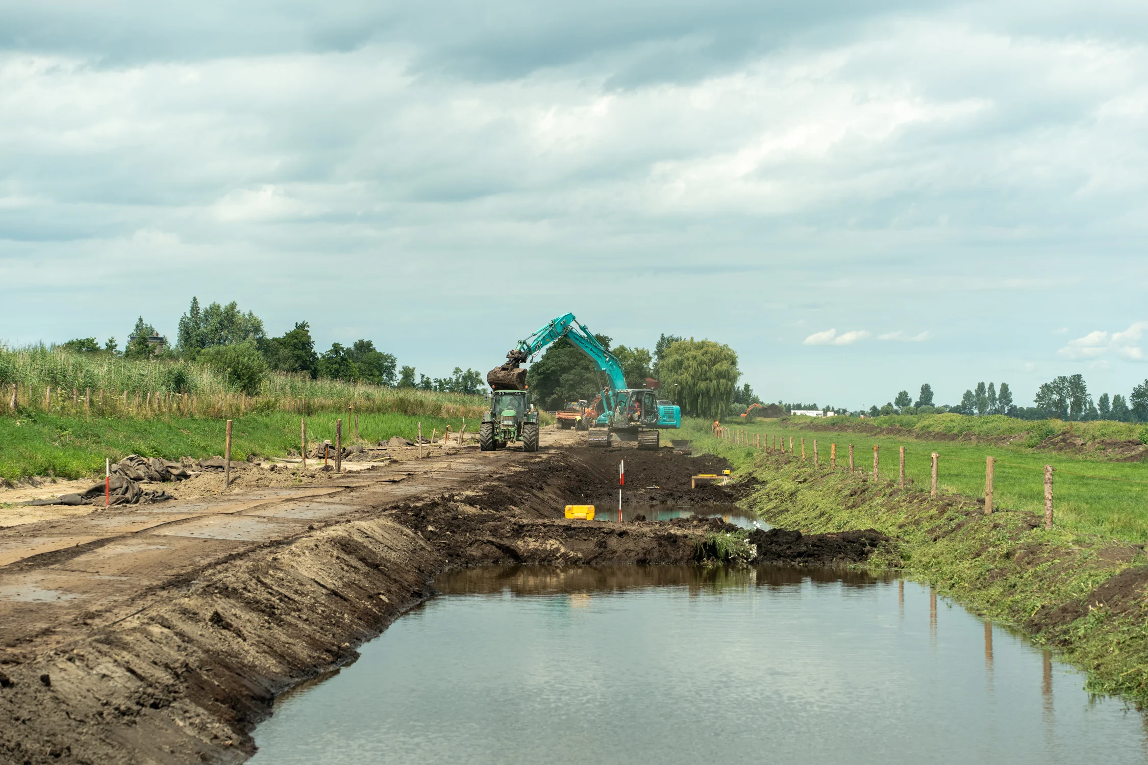Overzicht van de dijkverzwaringswerkzaamheden langs het water, met een kraan die grond laadt in een tractor op een langgerekt werktraject.