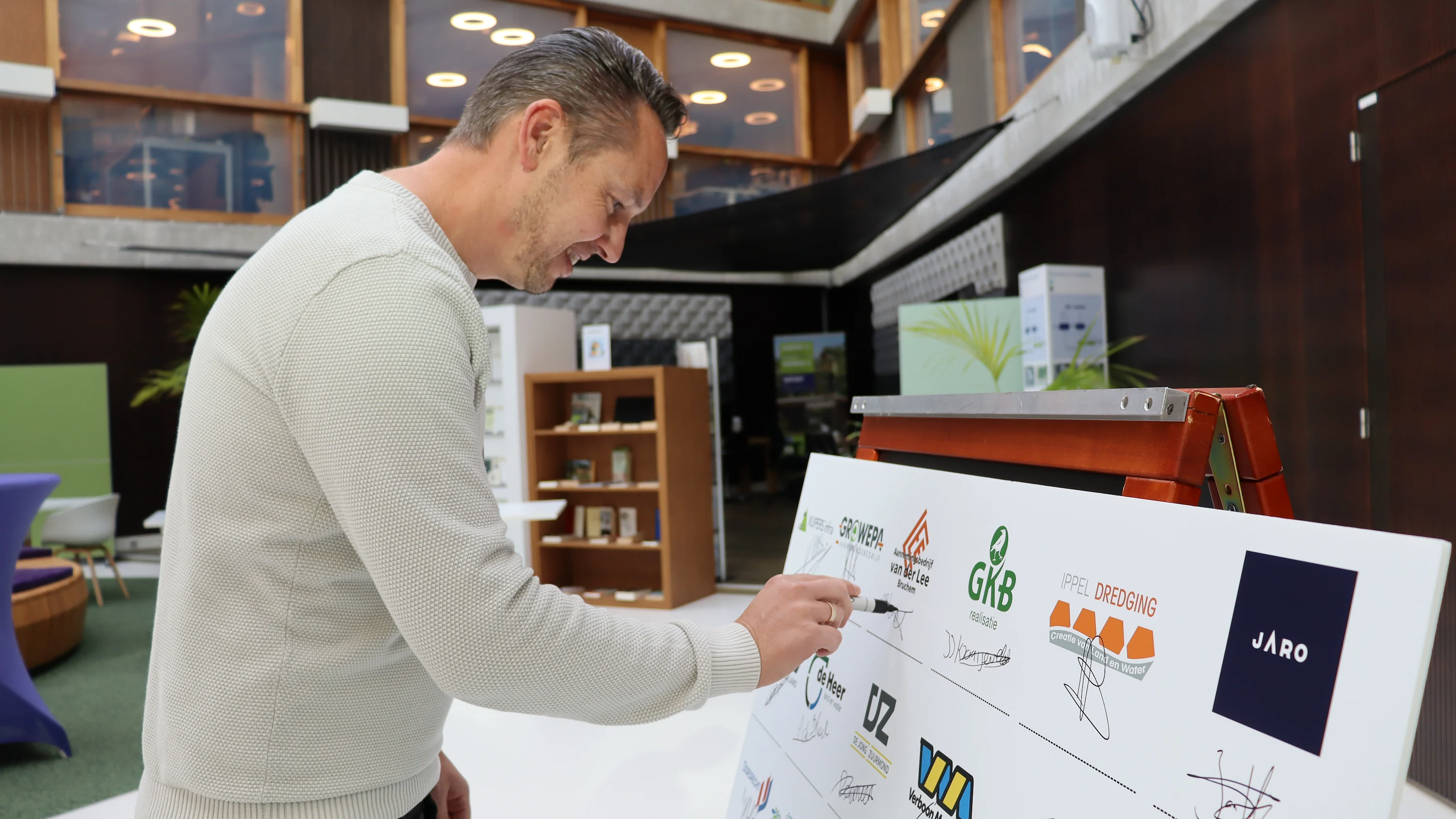 Een vertegenwoordiger van Van der Lee Bruchem zet zijn handtekening op het bord tijdens de officiële ondertekening van de Raamovereenkomst Stedelijk Baggeren, tussen de logo’s van deelnemende partijen.