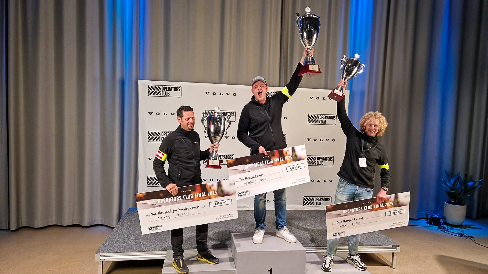 Prijsuitreiking van de Volvo Operators Club finale 2025: drie finalisten poseren op het podium met grote bekers en cheques, Arne Brals op de derde plek.