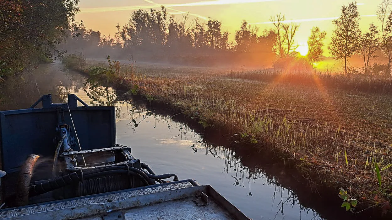 Zonsopkomst boven een veenlandschap met sloot, riet en nevelige bomen op de achtergrond, gezien vanaf een baggerboot.