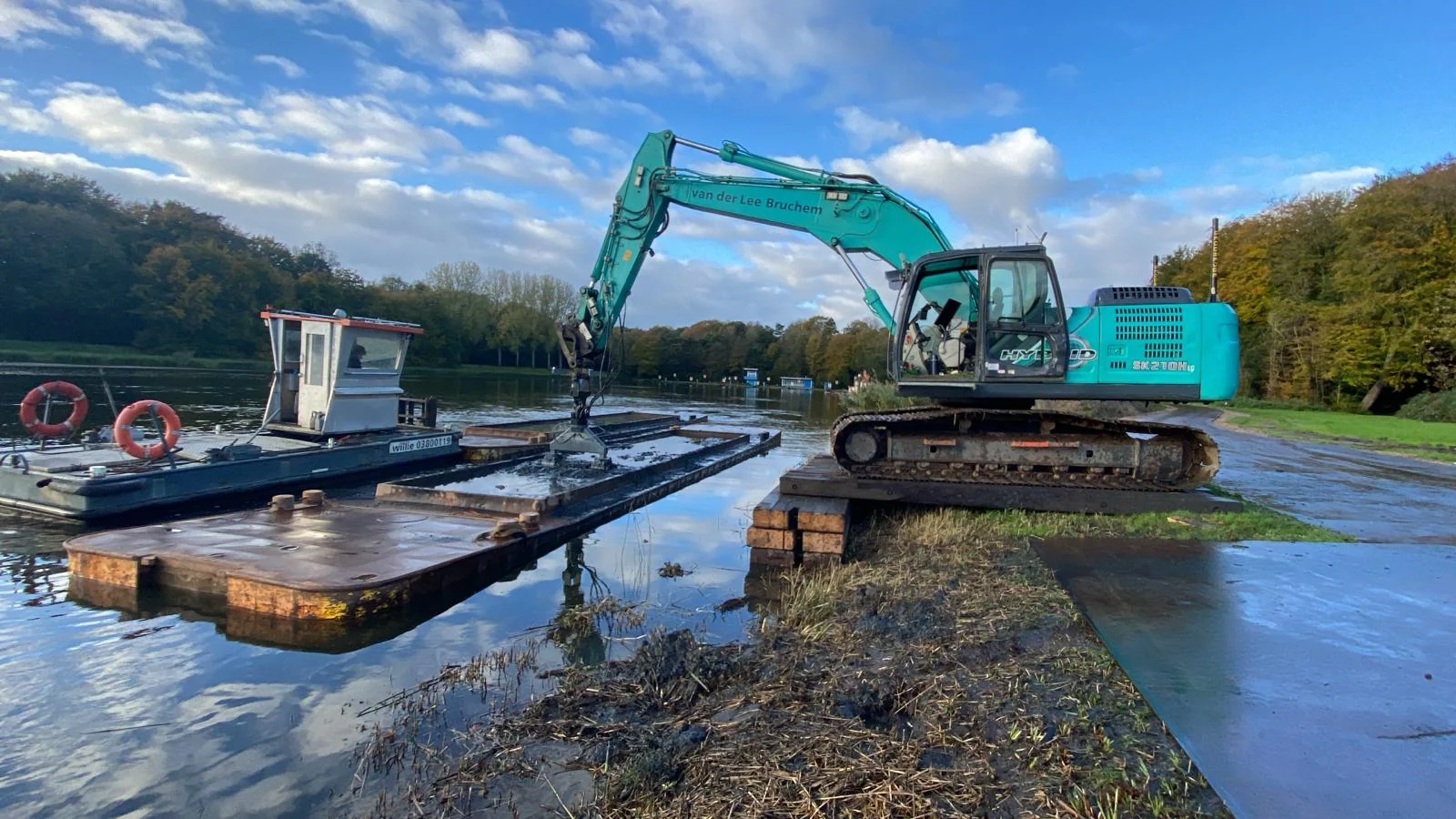 Een rupskraan van Van der Lee Bruchem aan het werk langs de waterkant van de Bosbaan, bezig met baggerwerkzaamheden vanaf een ponton.