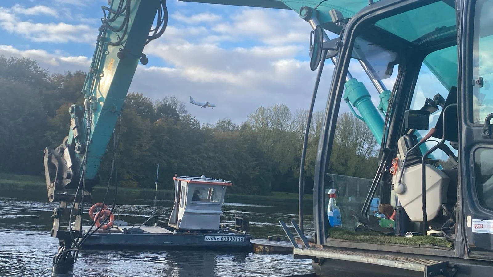 Zicht vanuit een graafmachine op de Bosbaan in Amsterdam, met een vliegtuig in de lucht op de achtergrond en een werkboot op het water.