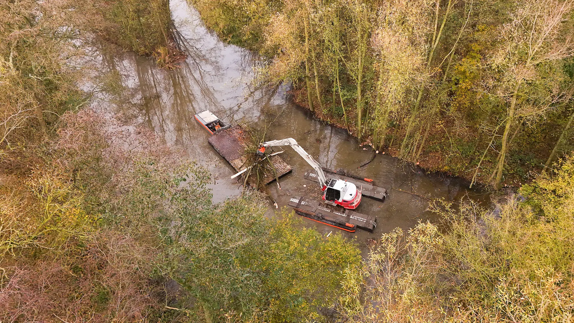 Een amfibische kraan van Van der Lee werkt midden in een smalle watergang, omringd door herfstige bomen. De kraan verwijdert takken en boomstammen uit het water.