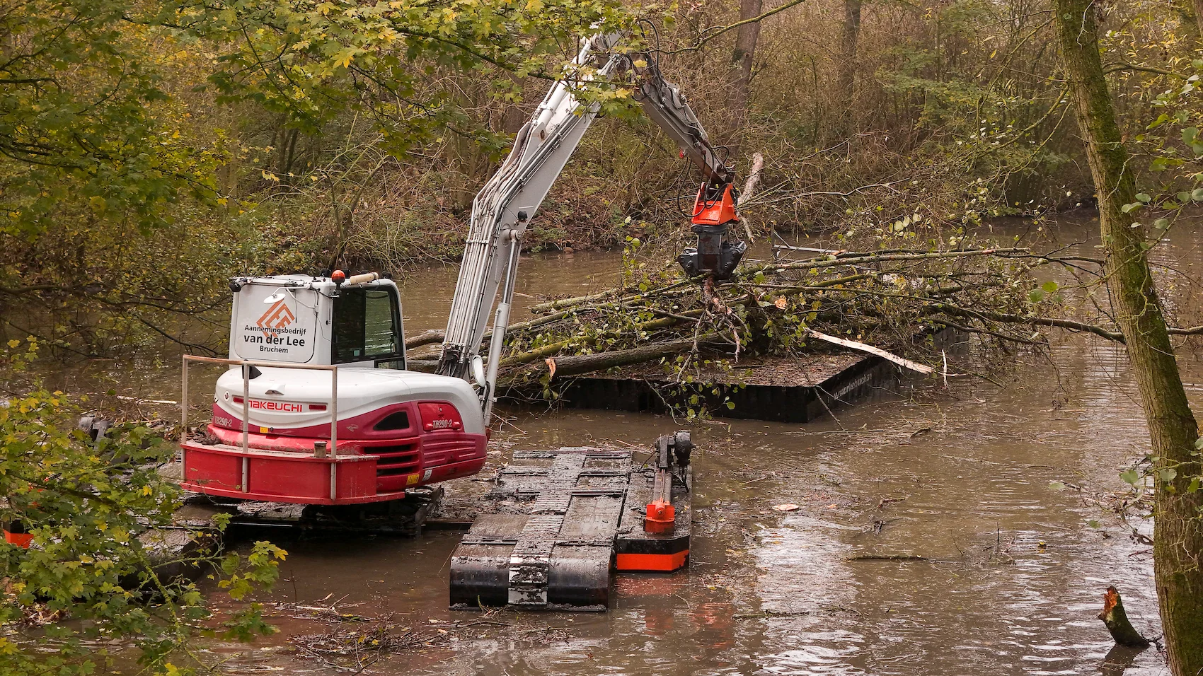 Een amfibische kraan van Van der Lee werkt midden in een smalle watergang, omringd door herfstige bomen. De kraan laadt boomresten op een ponton terwijl een medewerker meekijkt vanaf een werkschuit.
