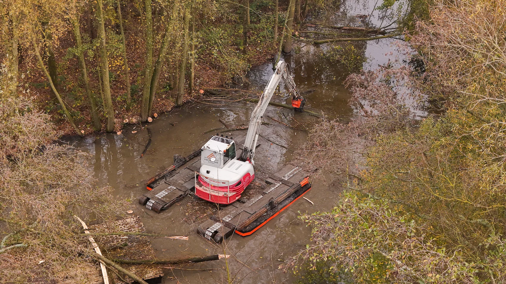 Een amfibische kraan van Van der Lee werkt midden in een smalle watergang, omringd door herfstige bomen. De kraan tilt een grote tak richting een ponton met werkschuit.