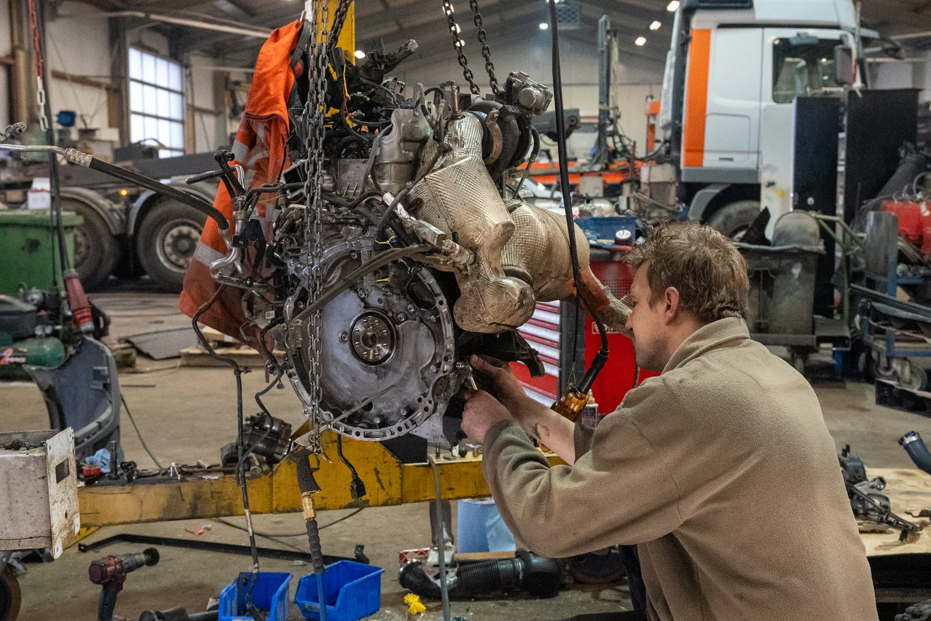 Een monteur werkt geconcentreerd aan een loshangende motor die met kettingen aan een hijsinstallatie hangt. De werkplaats is gevuld met trucks en gereedschap.
