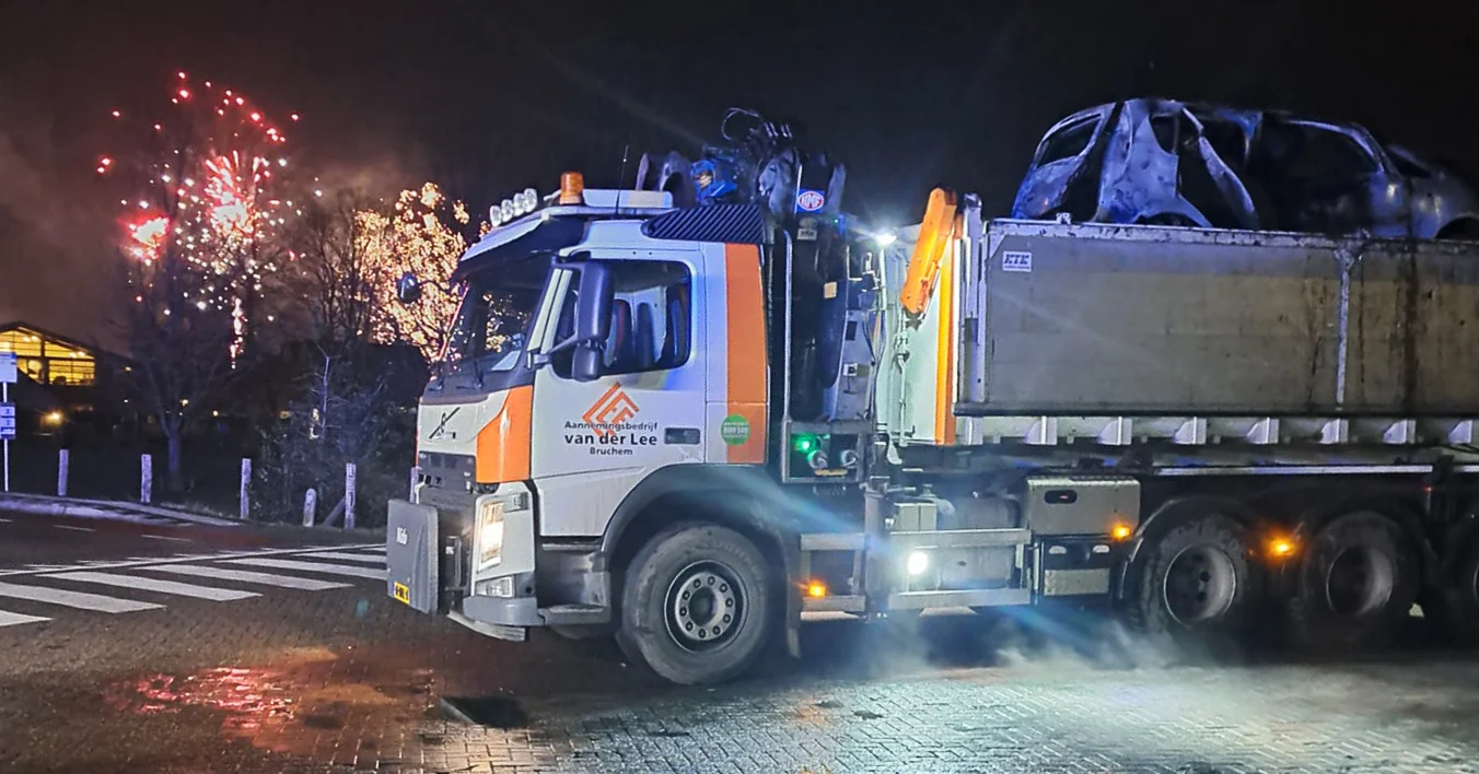 Knijperwagen van Van der Lee transporteert een uitgebrand autowrak, met vuurwerk in de lucht op de achtergrond