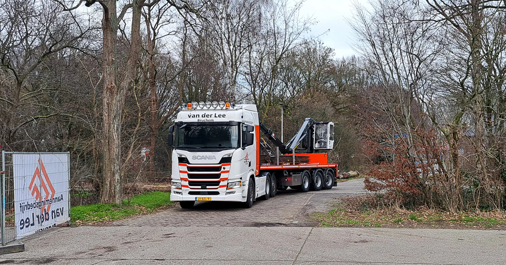 Witte vrachtwagencombinatie van Van der Lee Bruchem met nieuwe trailer en laadkraan, geparkeerd aan de rand van een bosrijk terrein bij een bouwhek met bedrijfslogo.