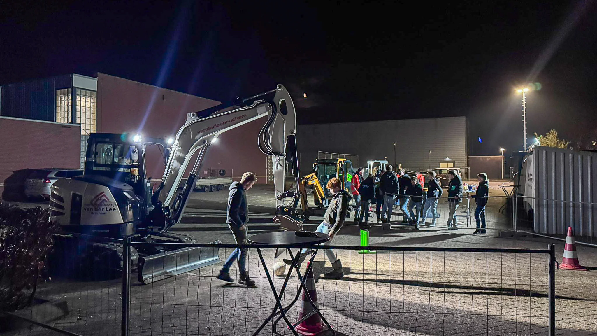 Mini graafmachine van Van der Lee Bruchem staat in de avond opgesteld op een verlicht terrein bij een oefenopstelling met pionnen en hekwerk