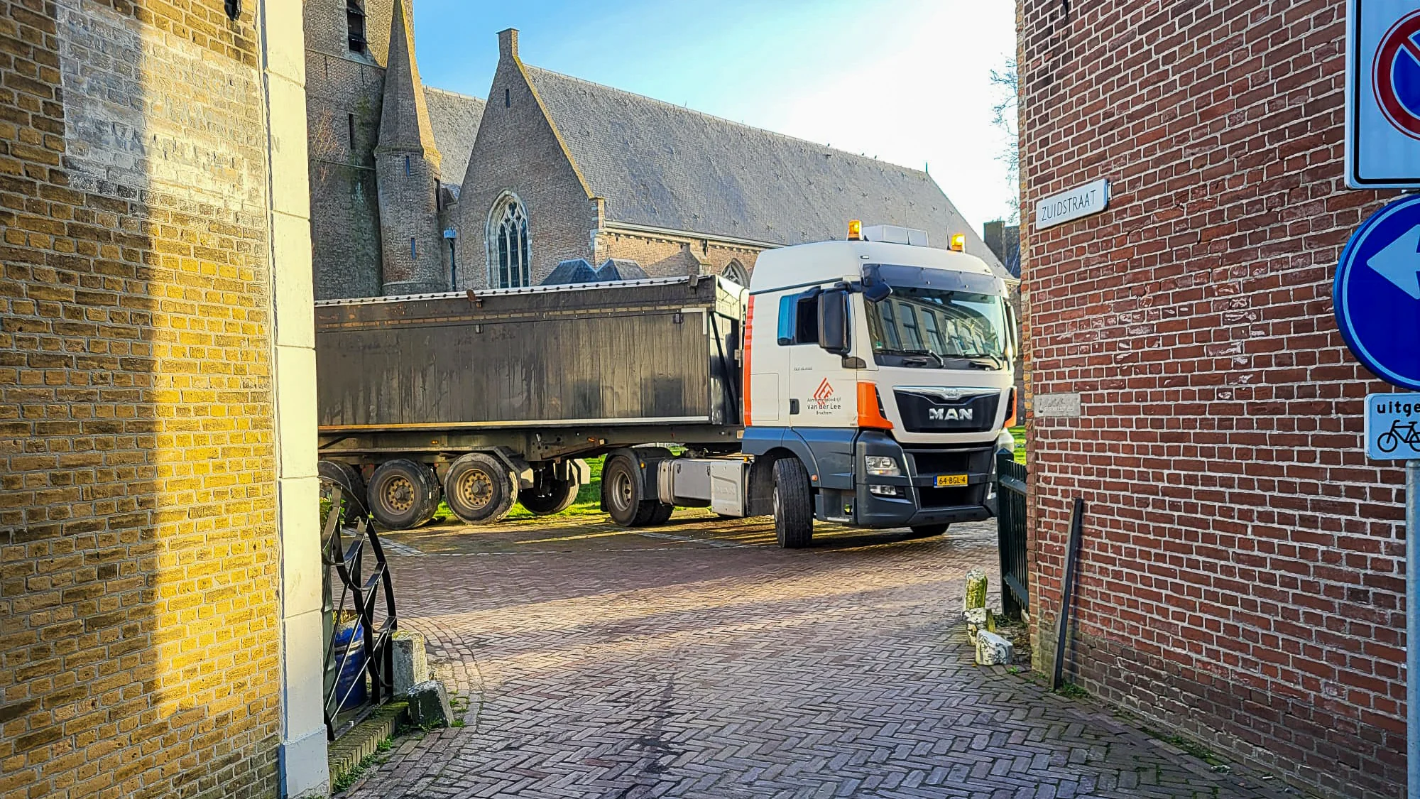Vrachtwagen van Van der Lee Bruchem manoeuvreert door een smalle straat naast een kerk in een dorpskern
