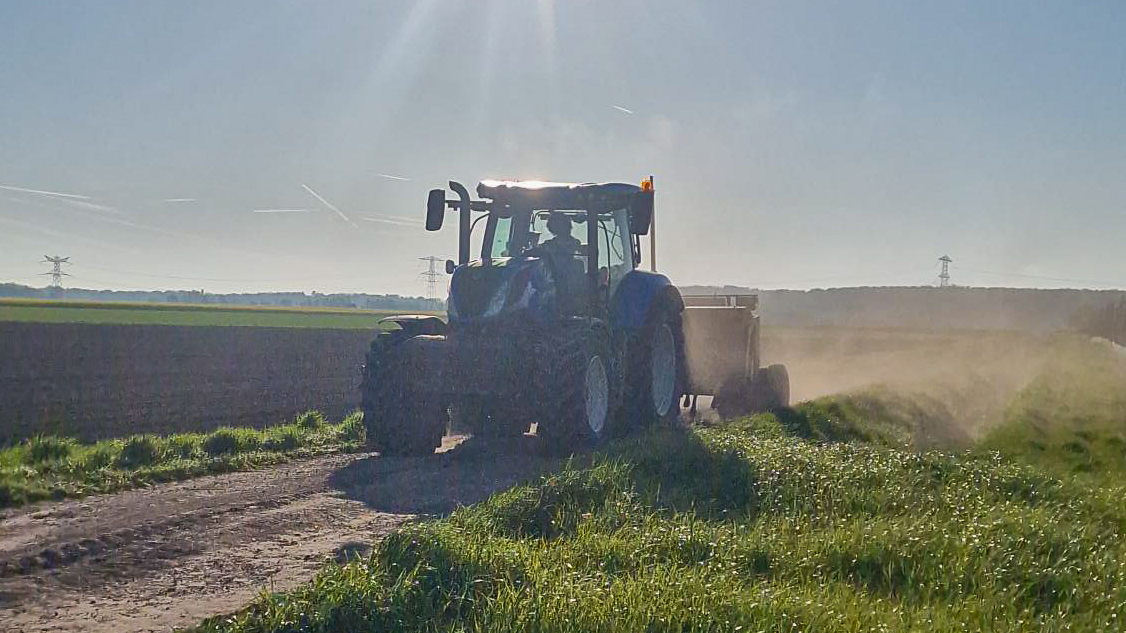 Tractor met dumper rijdt over een landelijke werkweg tijdens grondverzetwerkzaamheden bij zonsopkomst, met stofwolken zichtbaar achter het materieel.