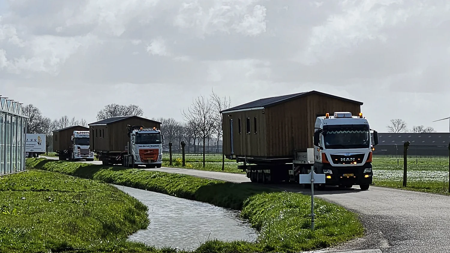Meerdere vrachtwagens vervoeren prefab chalets over een smalle landelijke weg tijdens transport voor [Houtenhuizen.nu](http://houtenhuizen.nu/).