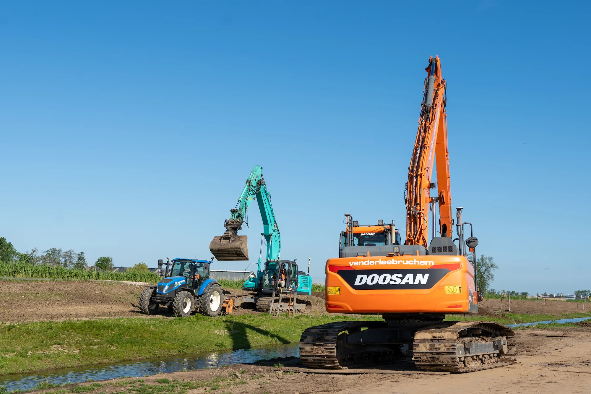 Graafmachines en tractor voeren grondverzetwerkzaamheden uit langs een watergang tijdens een infrastructureel project.