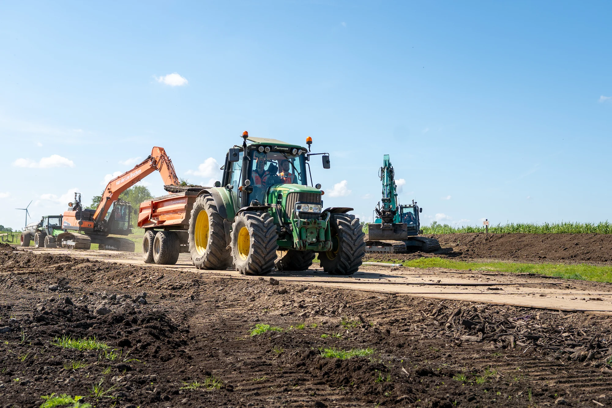 Tractor met dumper rijdt over de bouwweg terwijl graafmachines grondwerk uitvoeren langs een watergang.