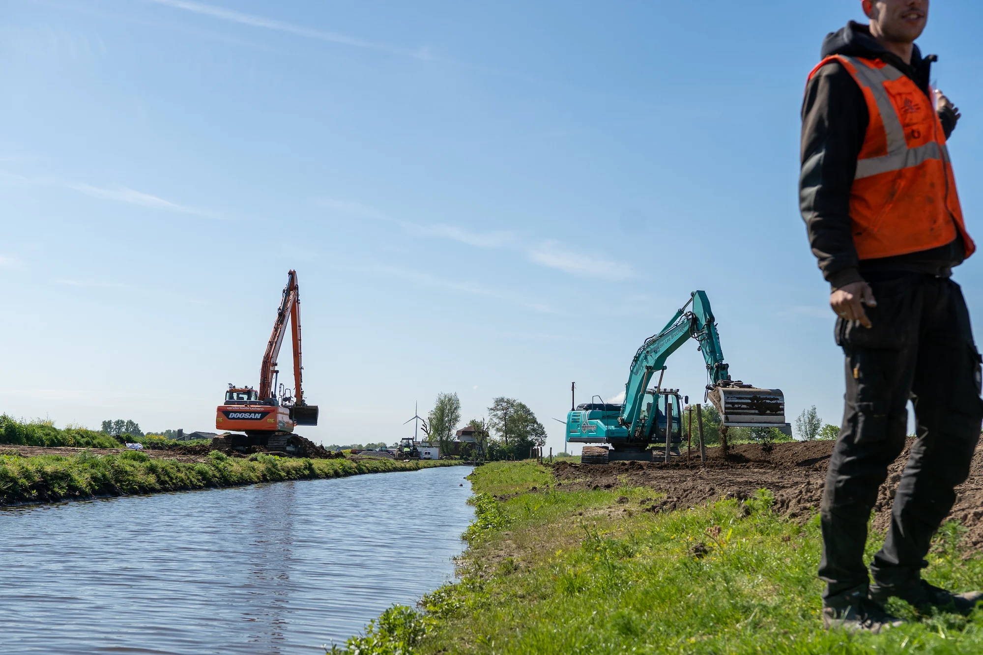 Overzicht van werkzaamheden aan een watergang met twee graafmachines en medewerker van Van der Lee Bruchem op de voorgrond.