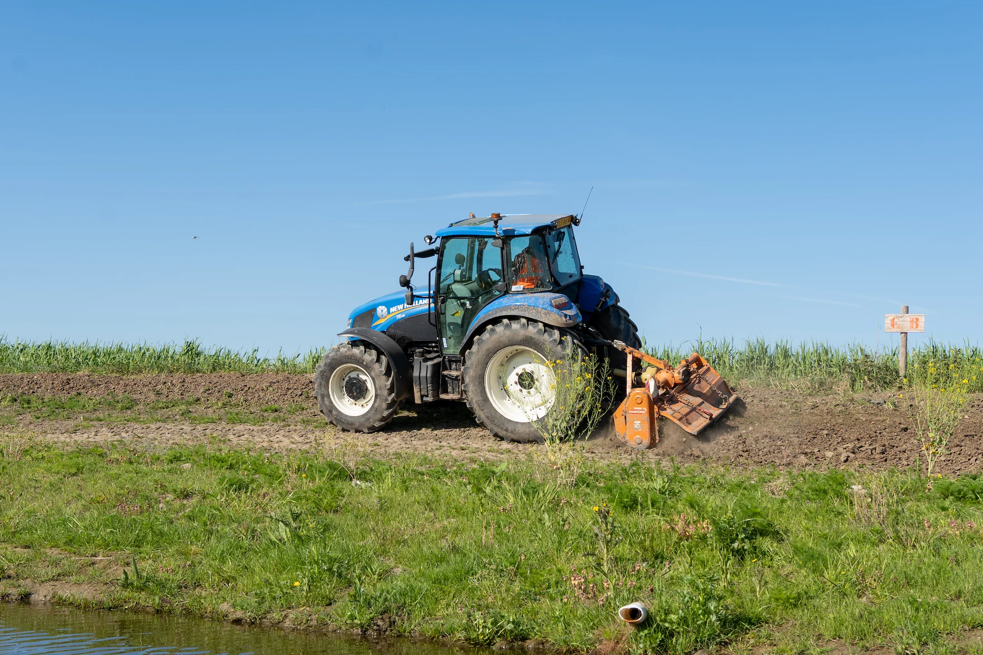 New Holland tractor voert frees en terreinonderhoud uit langs een watergang tijdens projectwerkzaamheden.