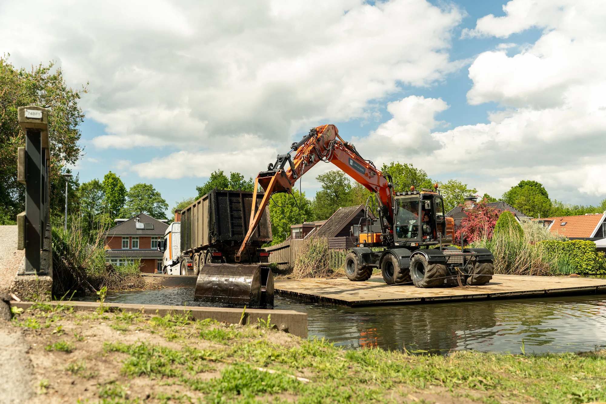 Mobiele kraan laadt bagger over vanuit de watergang naar een vrachtwagen op een tijdelijke werkvloer in een woonwijk.
