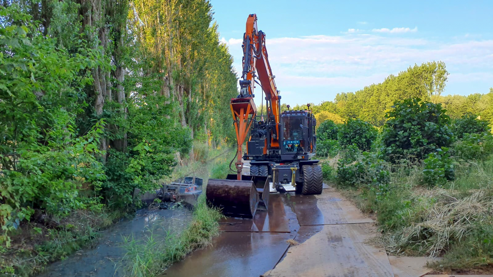 Mobiele kraan langs de watergang ondersteunt de werkzaamheden en werkt samen met de werkboot op een locatie met beperkte ruimte.