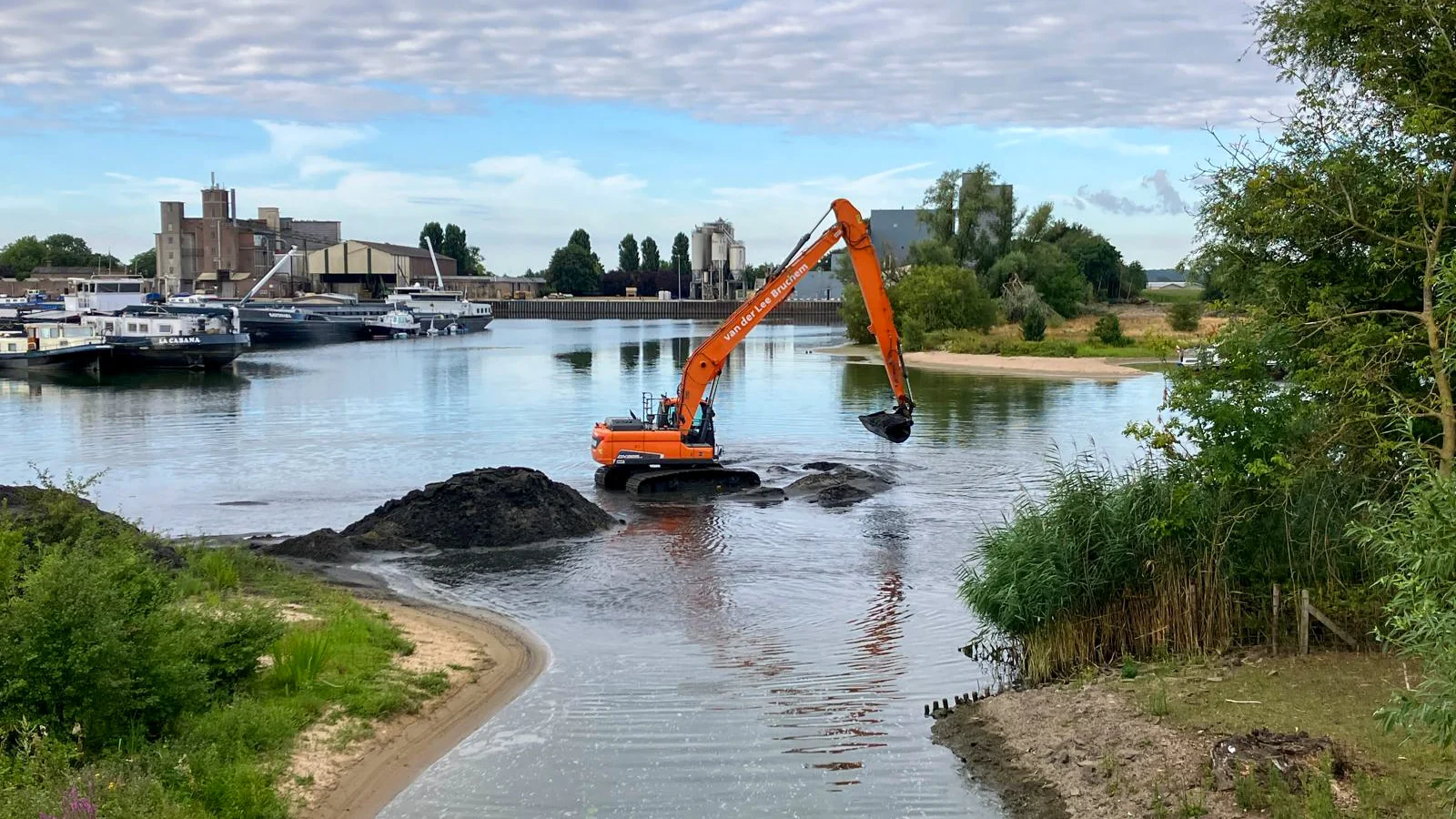 Breed overzicht van baggerwerkzaamheden waarbij een rupskraan van van der Lee Bruchem slib uit de watergang verwijdert. Op de voorgrond is de natuurlijke oever zichtbaar en op de achtergrond liggen diverse binnenvaartschepen aangemeerd.